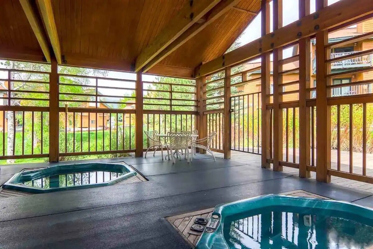 A covered area features two hot tubs, surrounded by wooden beams and a spacious floor. Natural light streams through the slatted wood walls, creating a bright and welcoming environment. A outdoor seating area with a table and chairs is visible in the background.