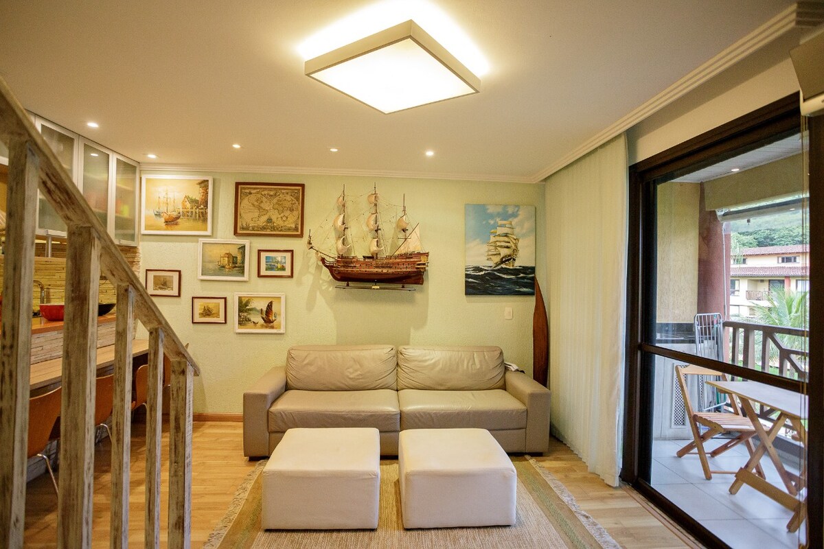 A living area is presented with a light-colored sofa and two white ottomans. Nautical-themed artwork and models adorn the walls, adding character. Natural light is allowed in through large sliding glass doors, leading to a balcony with outdoor seating.