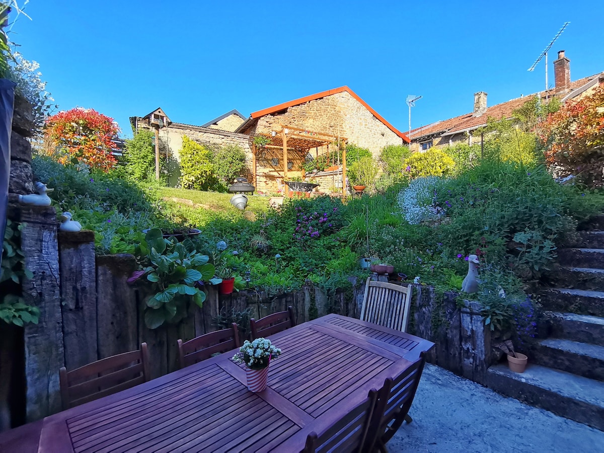 A spacious outdoor dining area is presented with a large wooden table surrounded by matching chairs. Lush greenery and colorful flowers create a vibrant backdrop leading to an inviting terrace and a stone house, framed by blue skies.