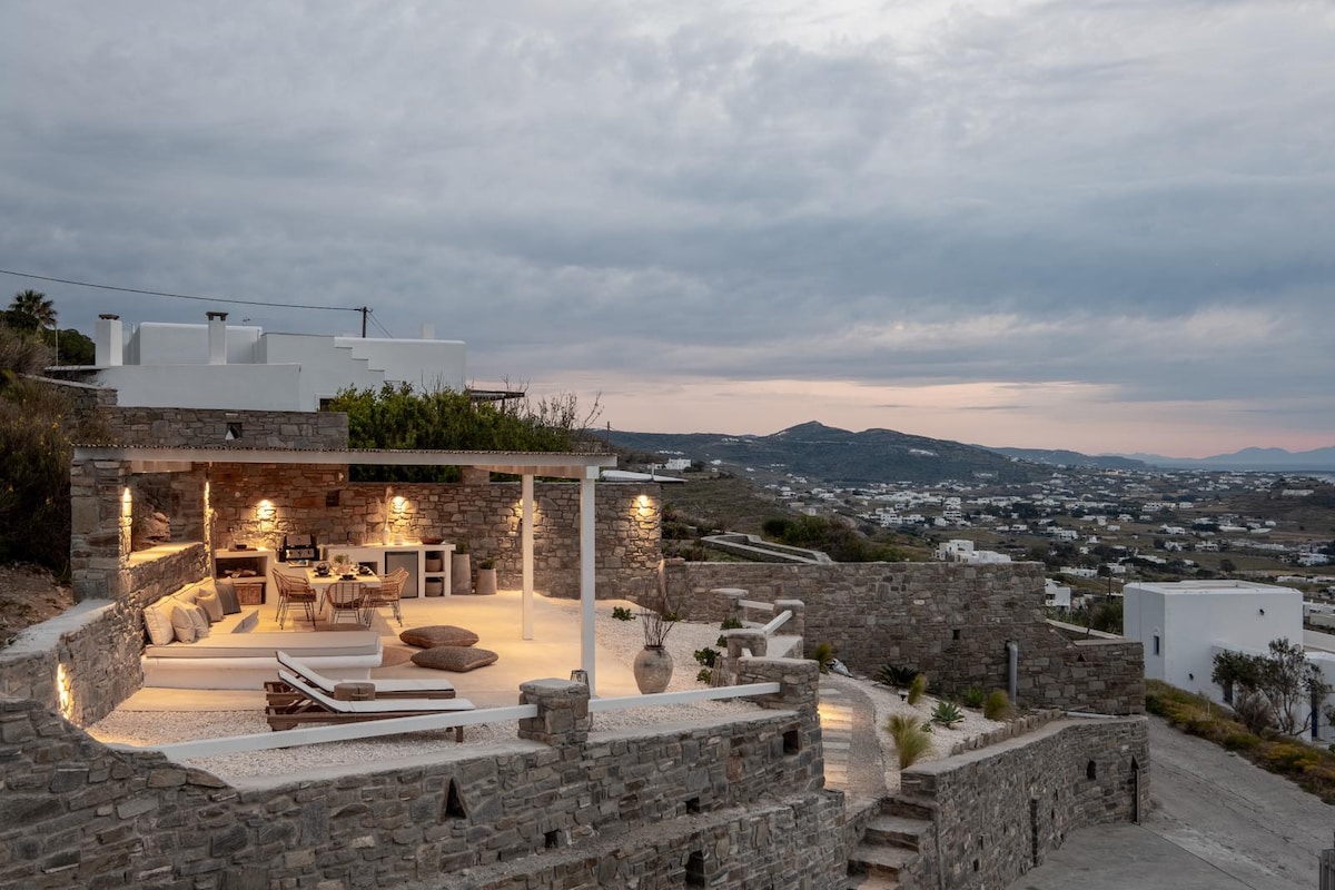An expansive outdoor terrace is depicted, featuring natural stone walls and stylish furniture. Soft lighting highlights the seating area and dining space, surrounded by scenic views of the landscape. The setting creates an inviting atmosphere for relaxation and gatherings.