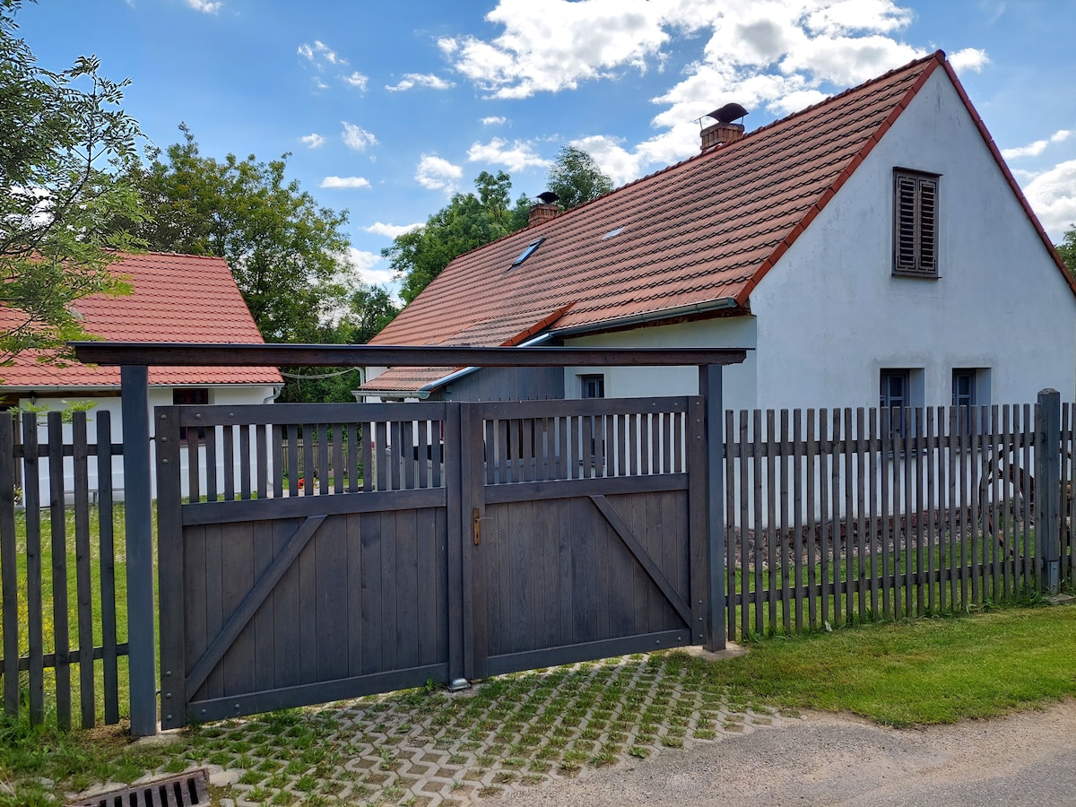 A wooden gate stands closed at the entrance to a property, framed by a gray fence. The cottage, with a sloped red roof and white exterior, is visible in the background alongside lush green grass and trees under a partly cloudy sky.