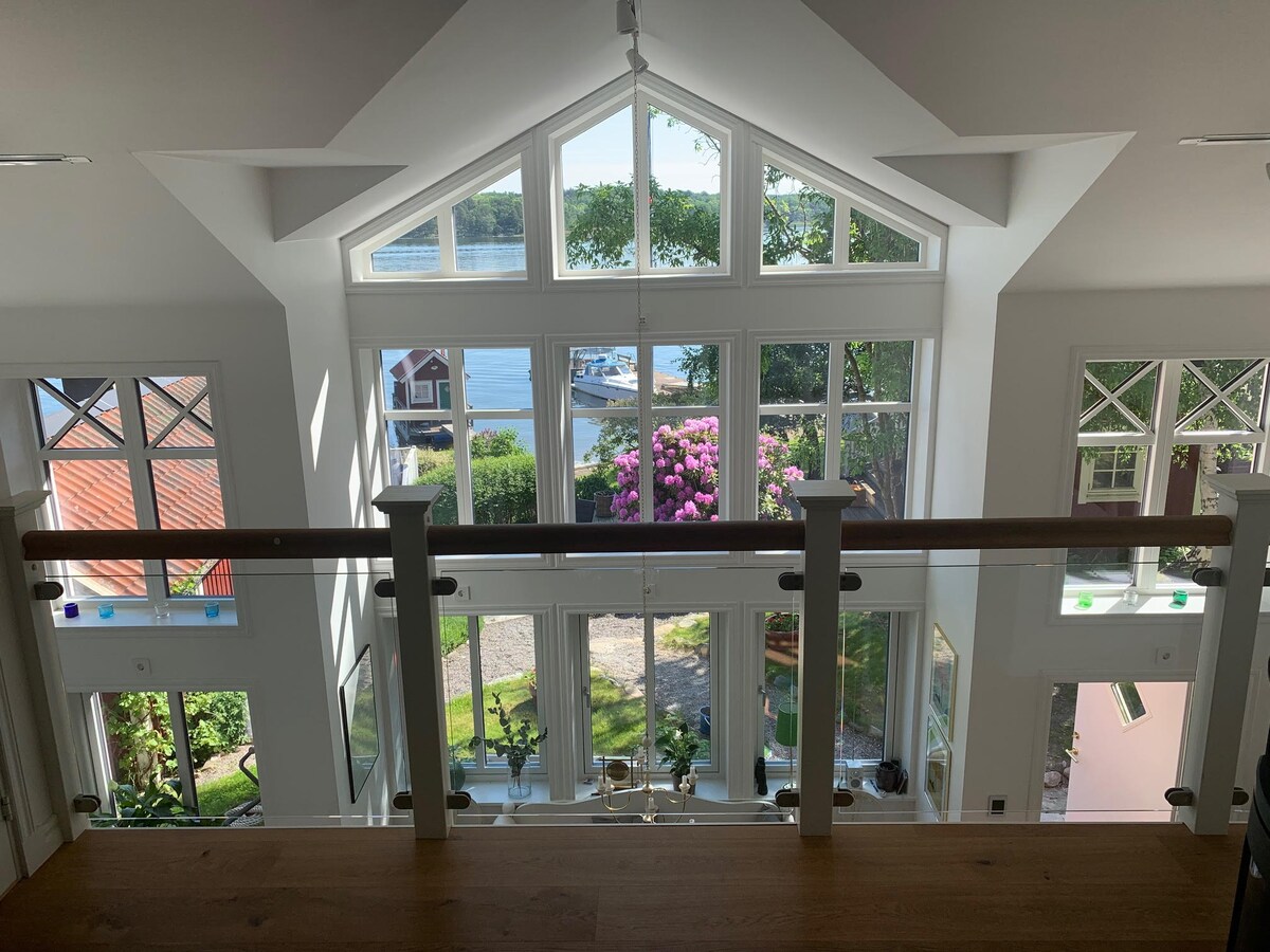 A spacious interior is visible from an upper level, featuring large windows that frame a view of the dock and lush greenery outside. Natural light floods the area, highlighting the modern architecture and decorative elements within the room.