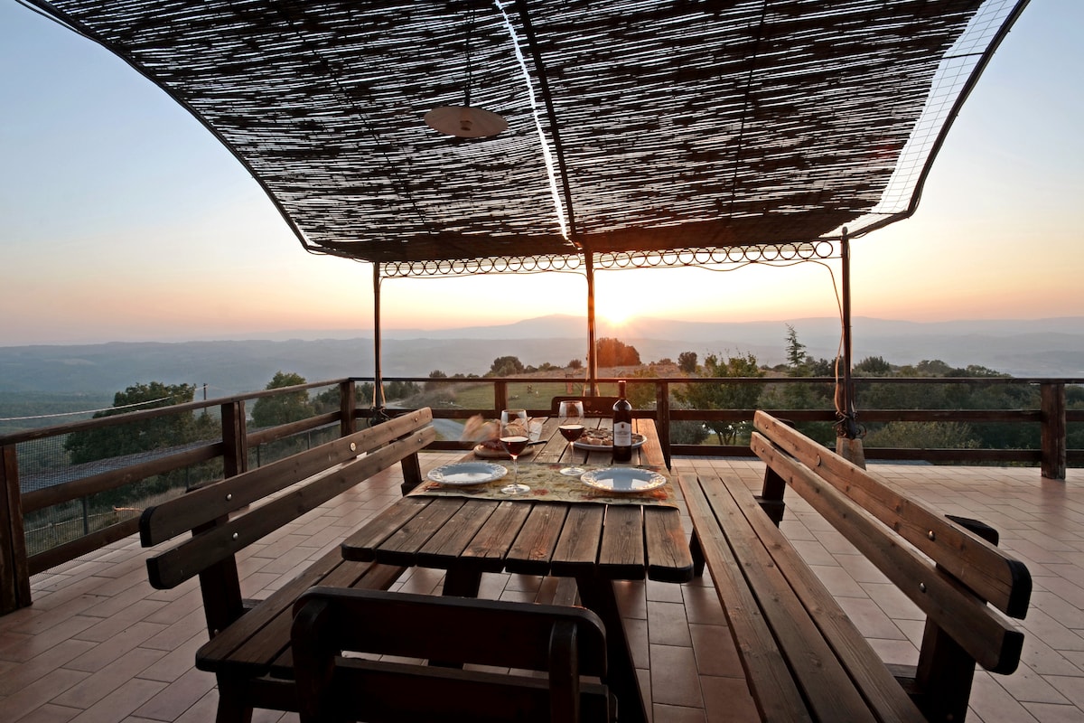 An expansive outdoor dining area features wooden tables and benches beneath a shaded pergola. Set against a backdrop of rolling hills, the scene captures a warm sunset, enhancing the serene atmosphere. Table settings include plates, glasses, and a bottle, inviting shared meals amidst nature.