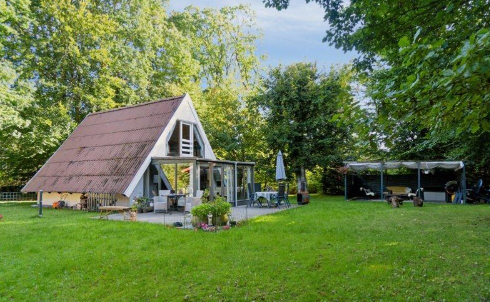 A charming summer house with a sloping roof is surrounded by lush green grass and tall trees. A covered pavilion is visible nearby, providing shaded seating. The patio area features comfortable furniture, and a small garden with blooming flowers enhances the inviting outdoor space.