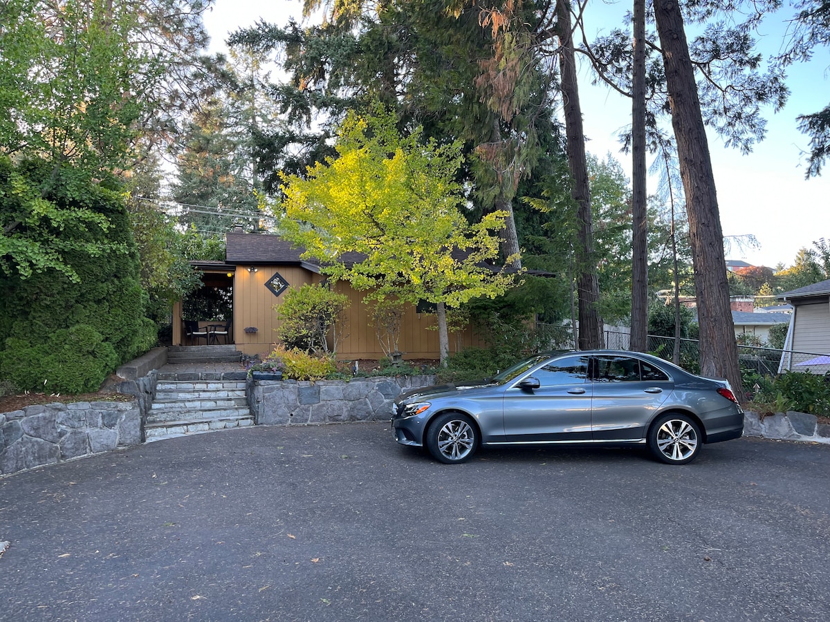 The exterior of Myrtle Cottage is shown, featuring a welcoming entrance set amidst lush greenery. A silver car is parked in front of the guesthouse, alongside neatly arranged stone steps leading to the main entry. Tall trees surround the property, enhancing its natural setting.