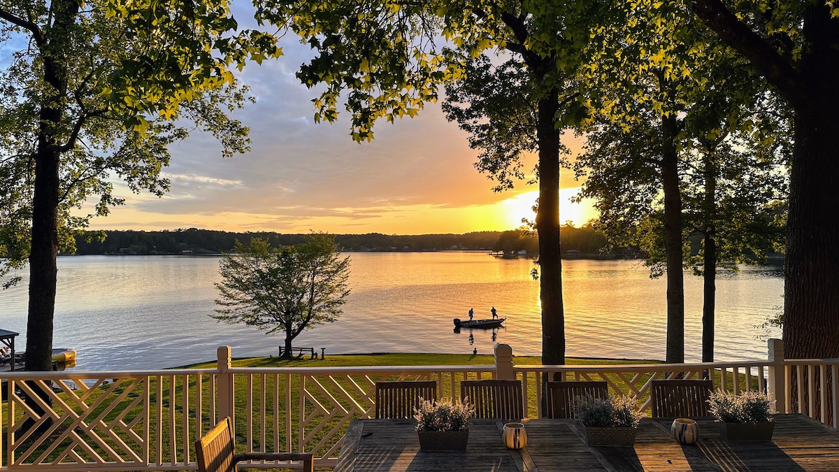 A scenic view of Lake Jackson is presented, showcasing a sunset casting golden hues across the water. Trees frame the scene, and a dining table with planters is visible in the foreground. A small boat is seen on the lake, enhancing the tranquil outdoor atmosphere.