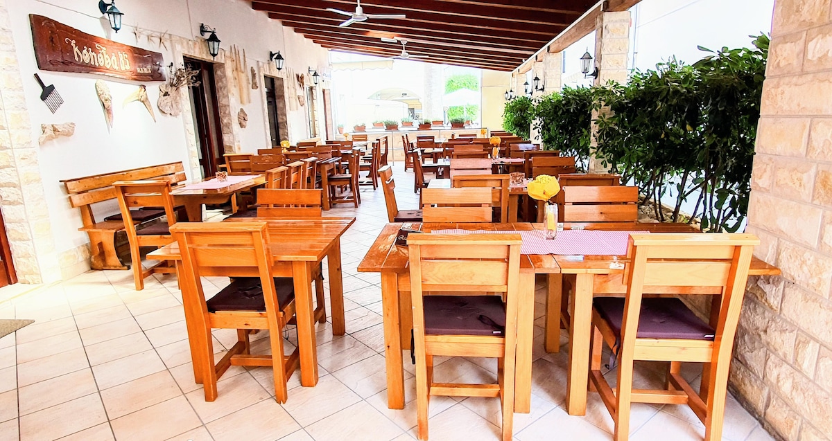 An outdoor dining area features wooden tables and chairs arranged neatly under a covered patio. The flooring is tiled, and decorative lanterns hang above, providing soft lighting. Lush greenery borders the space, creating a serene atmosphere for guests to enjoy meals al fresco.