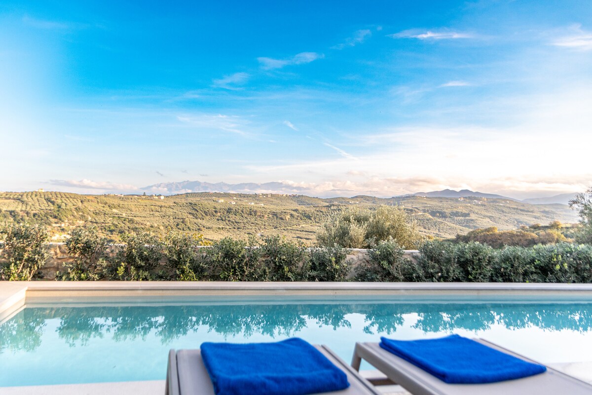 A private pool reflects the clear blue sky and lush hills, surrounded by greenery. Two lounge chairs with blue towels are positioned at the pool's edge, offering a serene spot to enjoy the picturesque mountain views.