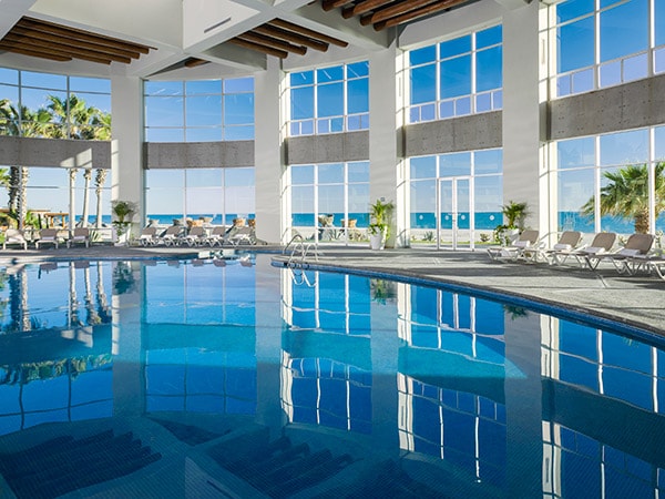 A spacious indoor pool area features large windows that fill the space with natural light, offering views of the exterior landscape. Comfortable lounge chairs are positioned around the pool, reflecting the tranquil blue water. Palm trees are visible beyond the glass, enhancing the serene atmosphere.