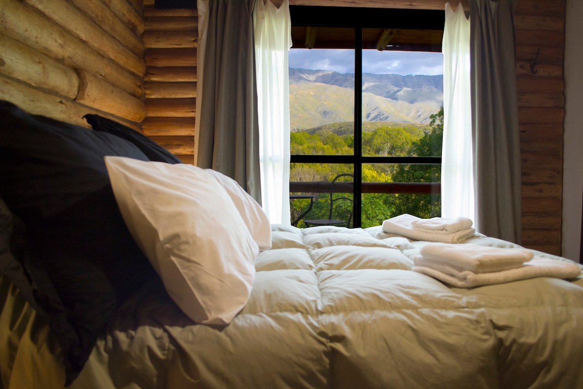 A spacious bed with crisp white linens is featured in this cozy room. Natural light pours in through a large window, offering a panoramic view of the surrounding mountains and greenery. Fresh towels are neatly arranged on the bed, enhancing the welcoming ambiance.