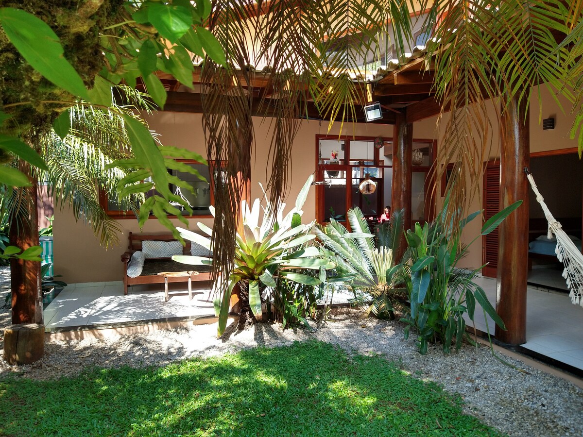 A lush garden area features diverse tropical plants surrounding a comfortable seating space. Hammocks can be seen hanging from wooden posts, providing a relaxed atmosphere. Natural light filters through the greenery, contributing to a serene and inviting outdoor setting.