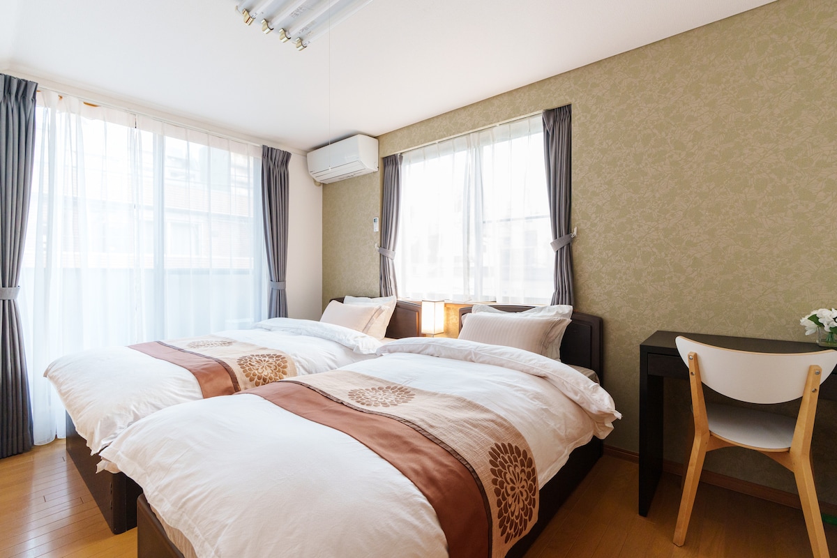 A bright guestroom features two spacious beds, each adorned with white bedding and terracotta accents. Natural light filters through the sheer curtains, illuminating the soft green wallpaper. A small desk and chair provide a functional workspace, enhancing the room's inviting atmosphere.