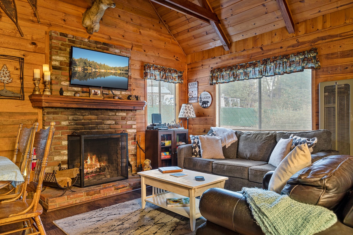 A cozy living room features a stone fireplace with a warm fire and a mounted television above. A large sectional sofa is positioned around a white coffee table. Natural light enters through two windows, and wooden beams enhance the vaulted ceiling.