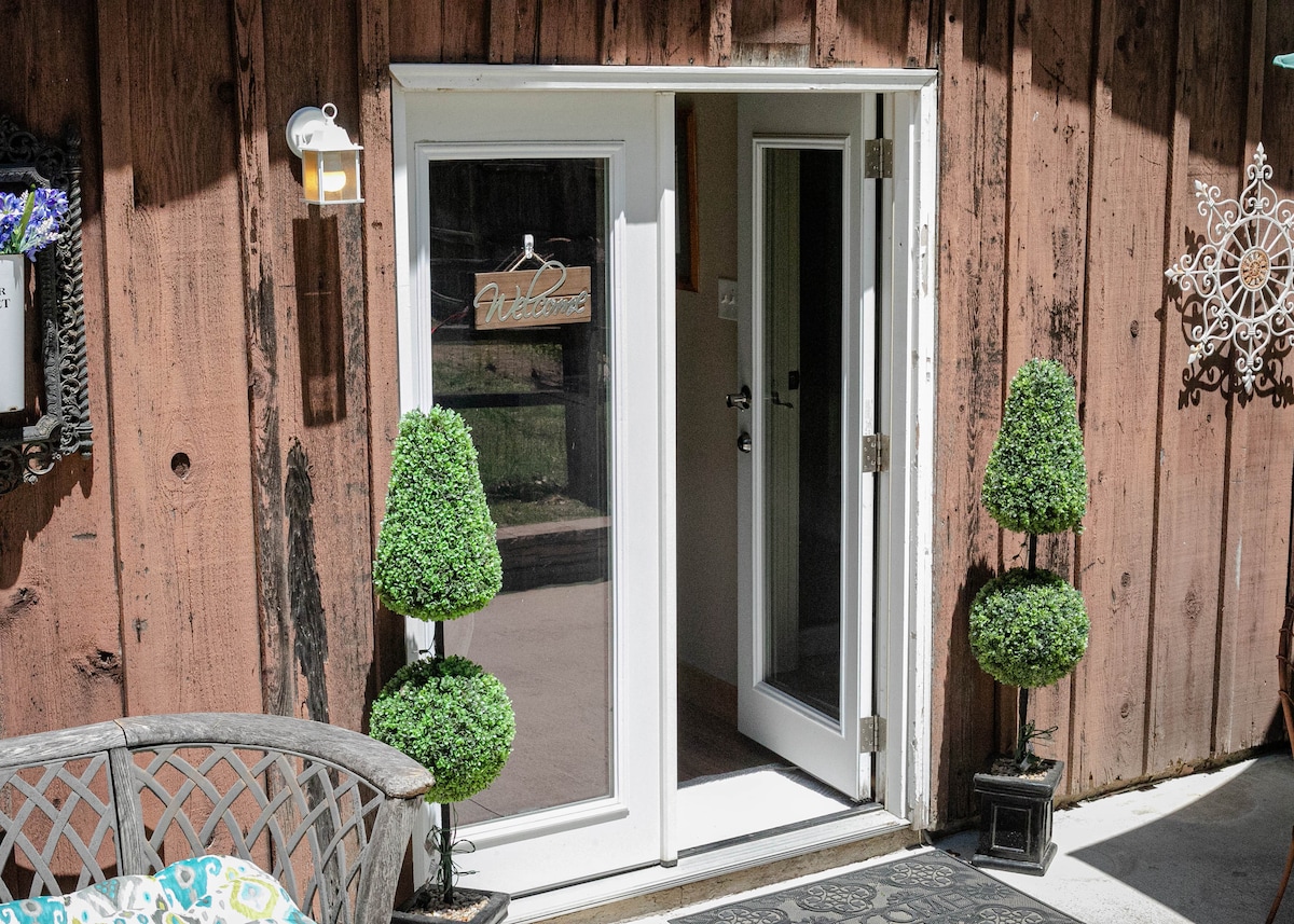 A welcoming set of double doors framed by tall, round topiary plants stands against a rustic wooden exterior. A small sign reading 'Welcome' hangs on one of the doors, adding a friendly touch to the entrance.