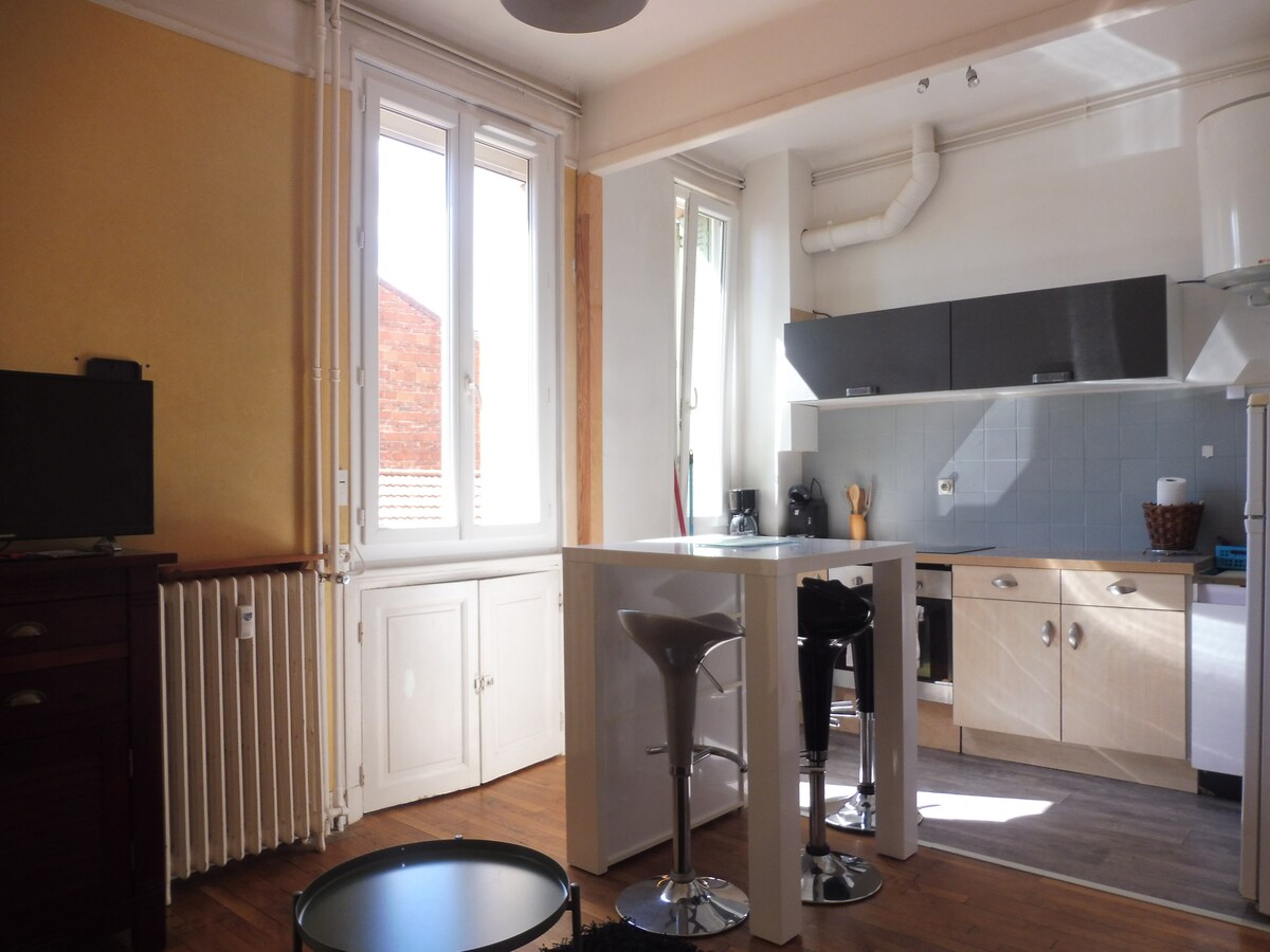 A compact kitchen area with light-colored cabinetry is visible, along with a modern bar table and stools. Large windows allow natural light to fill the space, highlighting the warm tones of the walls. A small television stands nearby, complementing the cozy ambiance.