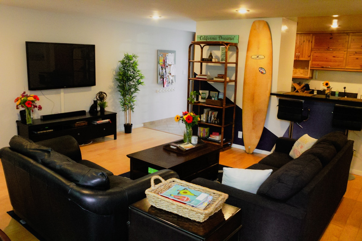 An open living area is depicted, featuring two seating areas with dark sofas and a central coffee table. A large television is mounted on the wall. A surfboard leans against the wall, and shelves hold decorative items, creating a relaxed atmosphere. A small dining area is visible in the background.