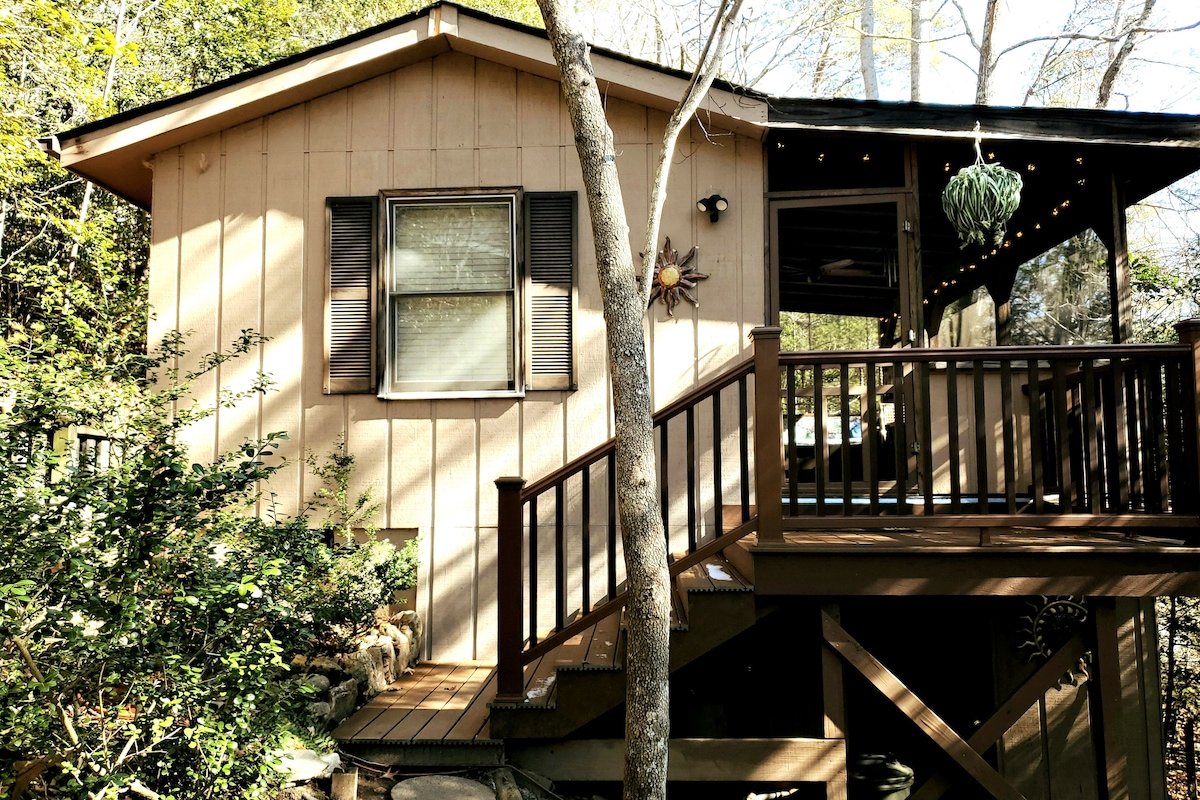 The cabin exterior is surrounded by trees, featuring light brown siding and a welcoming deck. Two windows are adorned with shutters, and a small decorative sun element is visible on the wall. A set of stairs leads up to the entrance, enhancing access to the cabin.
