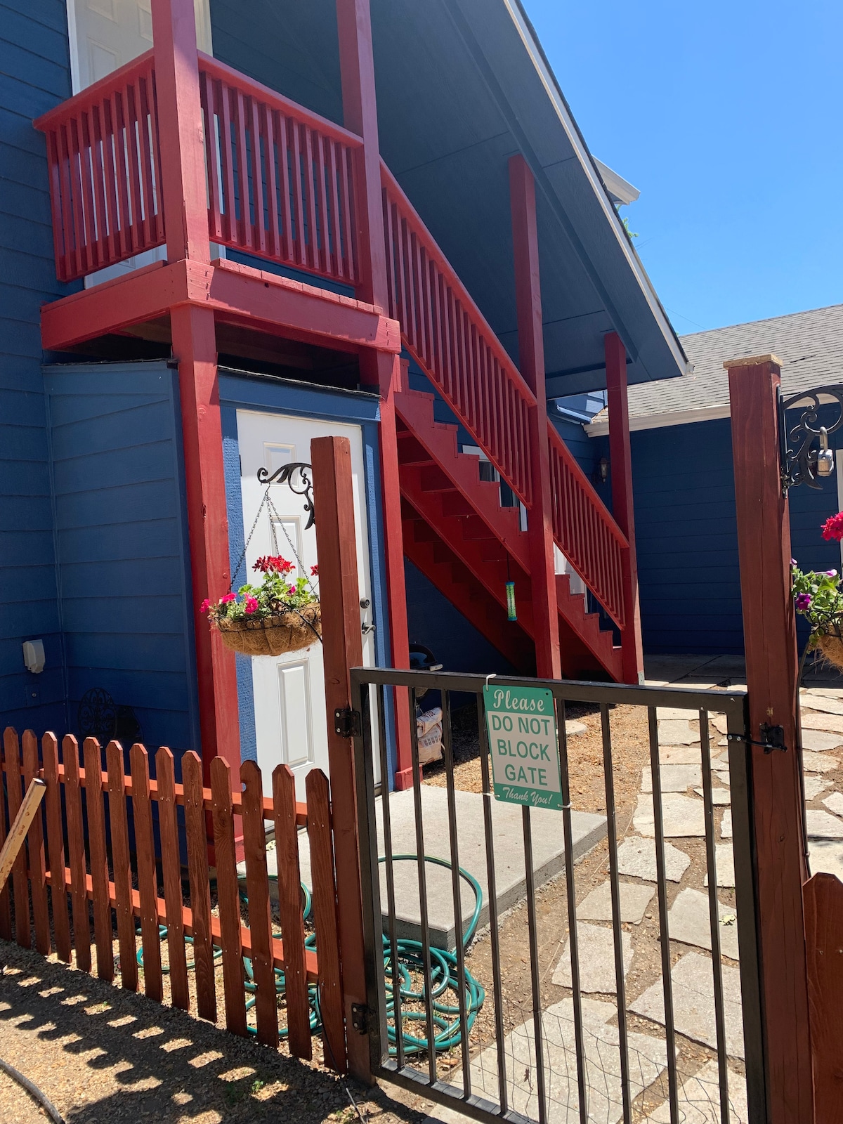 A vibrant entrance is framed by a wooden fence and gate, leading to the stairs of the second-floor studio. Flower baskets hang from the porch, complementing the blue exterior. Clear blue skies provide a bright backdrop to the welcoming atmosphere.