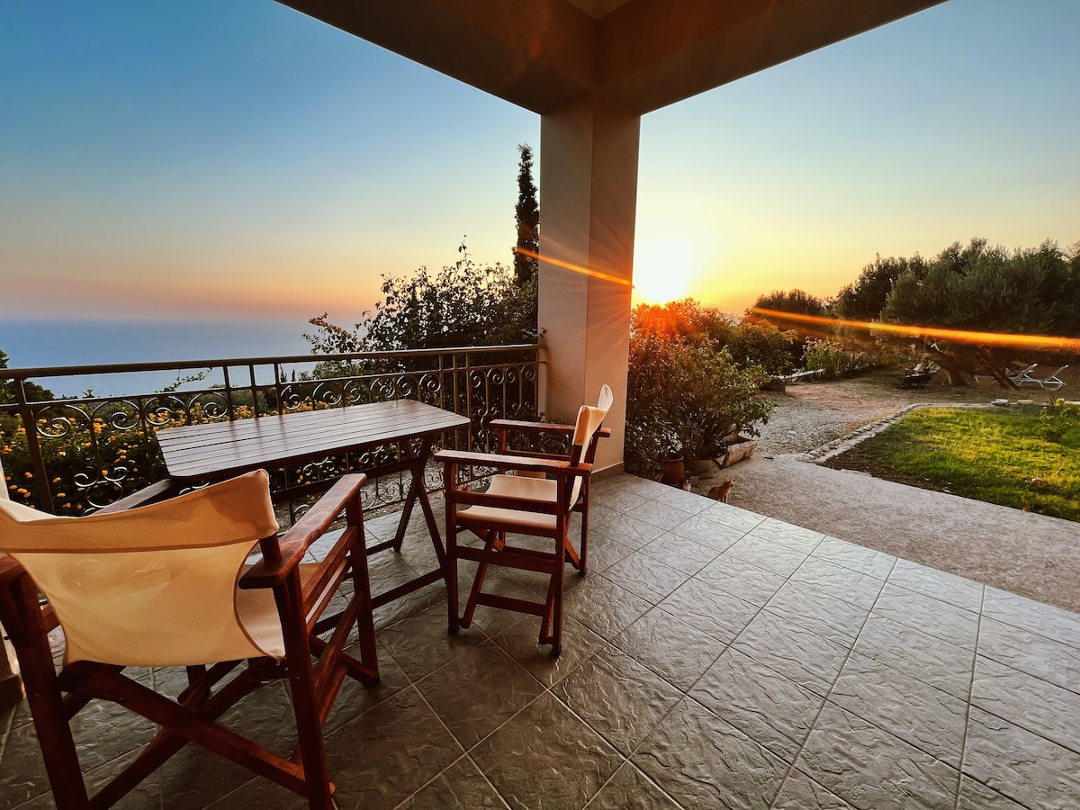 A shaded patio features a wooden table and two folding chairs, positioned to enjoy the sunset. The view extends towards the garden, where olive trees and decorative plants can be seen. The expansive sky transitions into warm hues, creating a serene atmosphere.