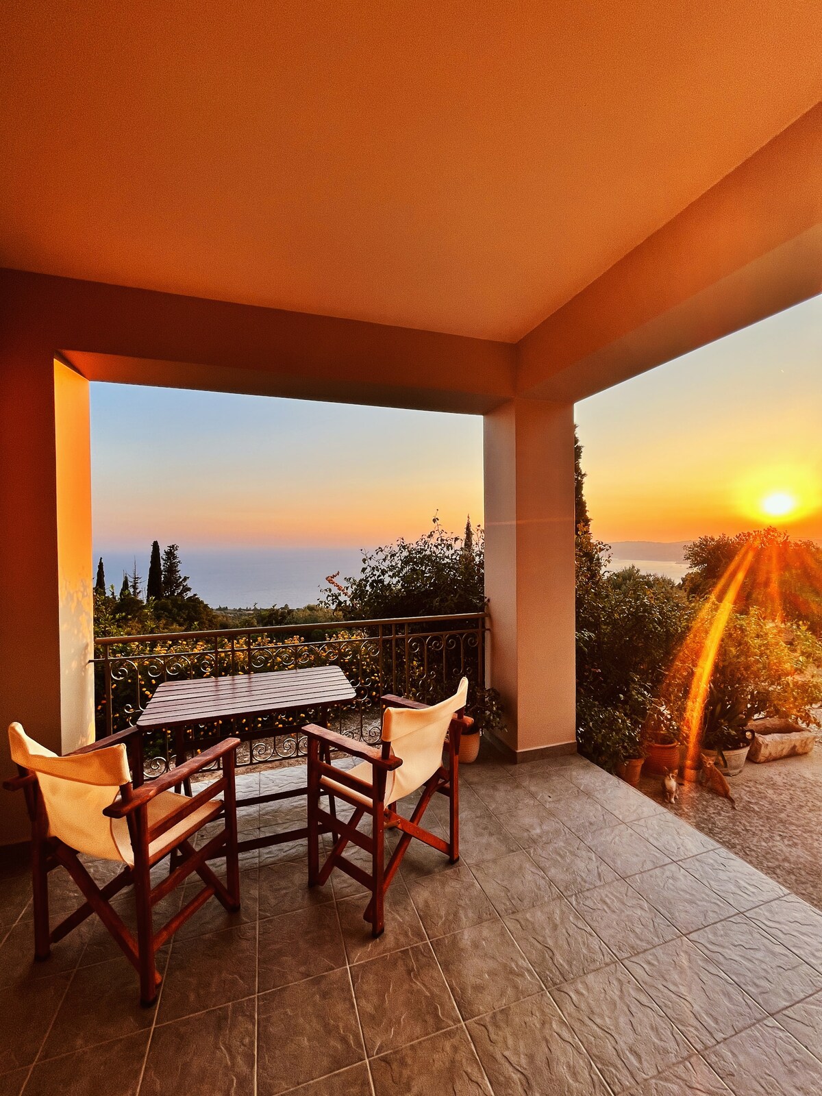 A spacious patio area features a wooden table set with two chairs, framed by an open archway. The view showcases a sunset over the horizon, with warm golden light illuminating the surrounding greenery.