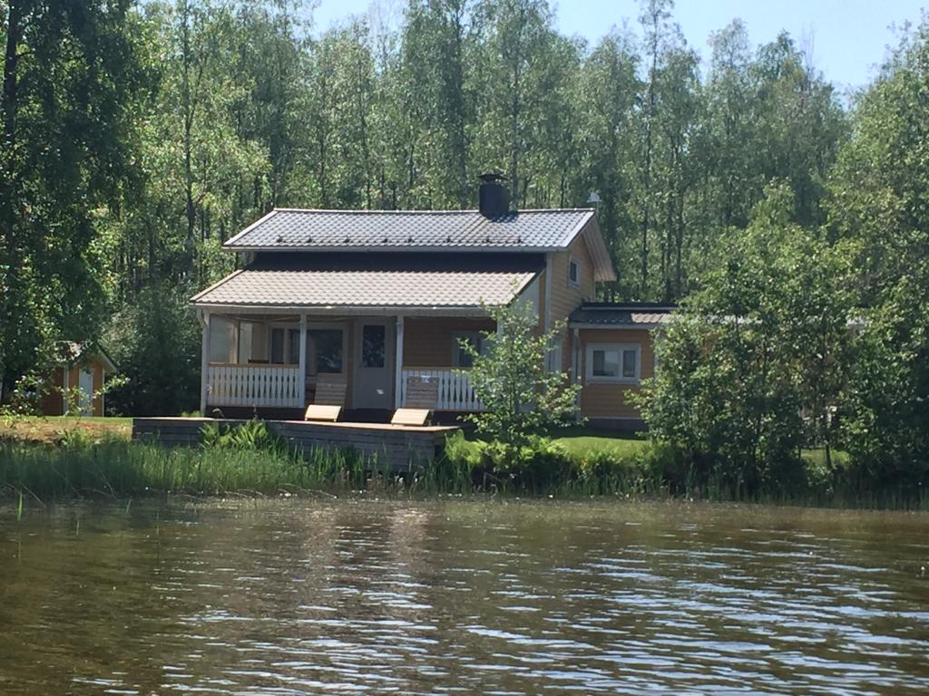 A wooden cottage stands by the lakeshore, surrounded by trees and greenery. Its two-story structure features a covered porch with seating and a spacious roof. A small dock can be seen nearby, providing access to the clear water of the lake.