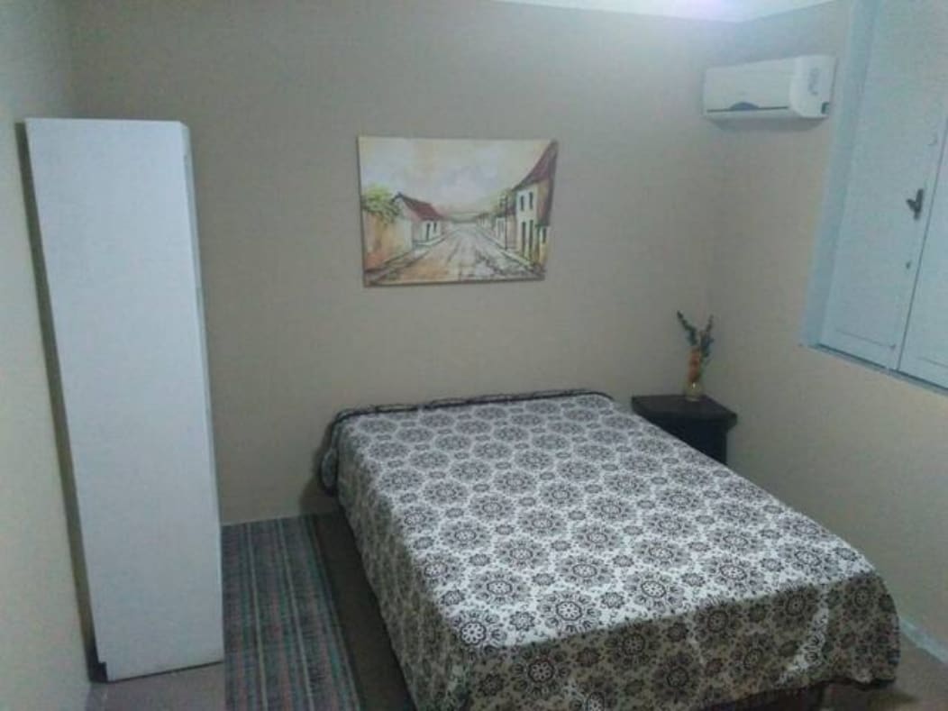 A well-appointed sleeping area features a spacious bed covered with a patterned blanket. A nightstand is positioned beside the bed, and a decorative piece is placed on it. A window with blinds allows light to enter, while a standing wardrobe provides additional storage.