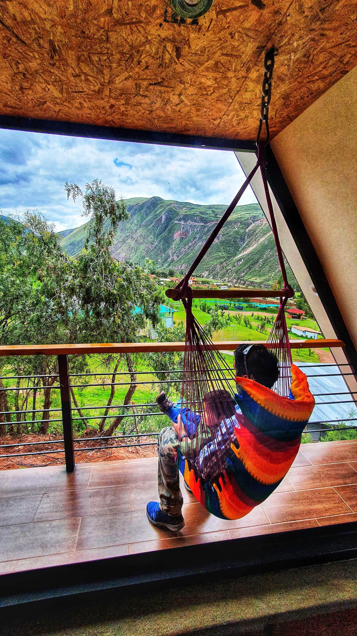 A colorful hammock is suspended from the ceiling, providing a cozy spot to relax. The view reveals lush green valleys and mountains under a bright sky, creating a serene backdrop. Natural light floods the space through a large window, enhancing the connection with nature.