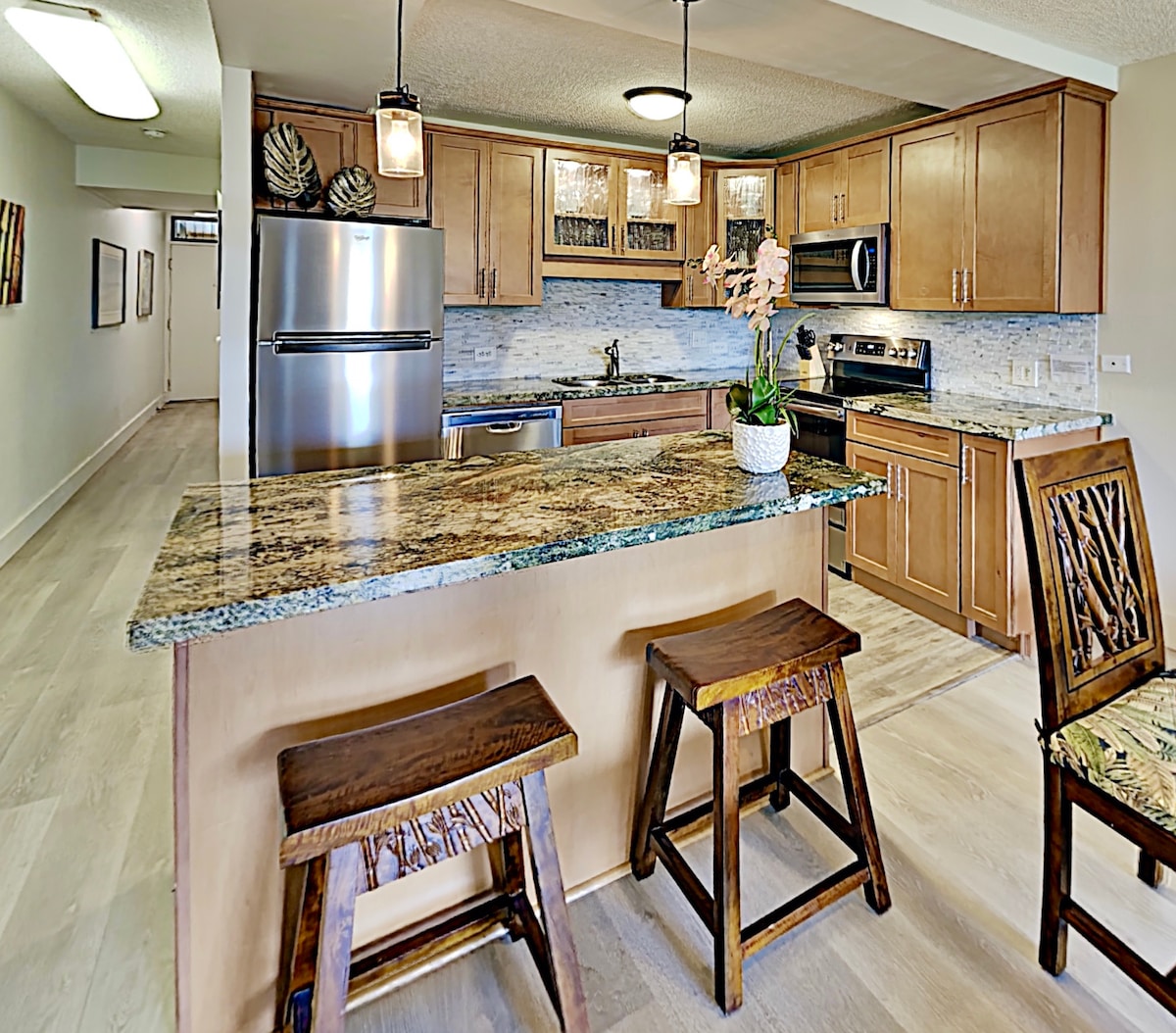 The kitchen features modern appliances, including a stainless steel refrigerator, stove, and microwave. A spacious island with a granite countertop is accompanied by three wooden stools. Natural light illuminates the elegant cabinetry and decorative tile backsplash, creating a welcoming cooking space.