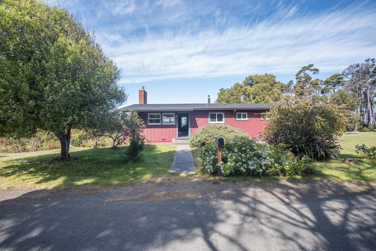 A single-story home with a red exterior is set amidst a grassy landscape, surrounded by various shrubs and trees. A clear path leads to the front door, which is flanked by a small outdoor area featuring flowering plants.