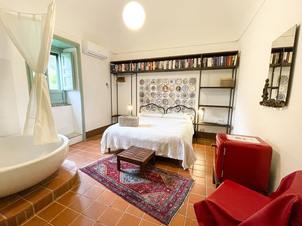 A spacious bedroom features a comfortable bed adorned with a white quilt. Shelves above the bed display decorative plates and books. A cozy seating area with a red chair complements the room, which includes a round bathtub and an air conditioning unit. Natural light streams through a window.