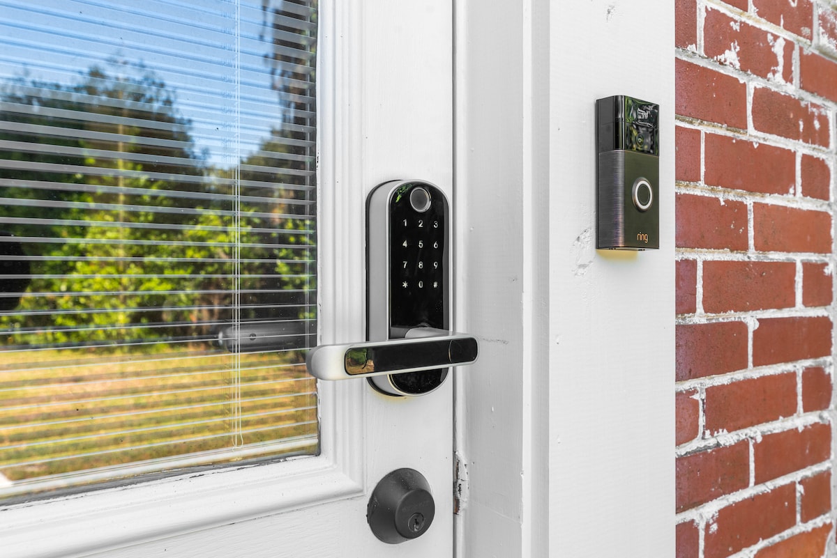 The entrance features a secure electronic keypad lock and a video doorbell. Natural light filters through glass panels beside the door, illuminating the white frame and red brick wall. A green lawn can be seen beyond the entrance, adding to the overall welcoming aspect.