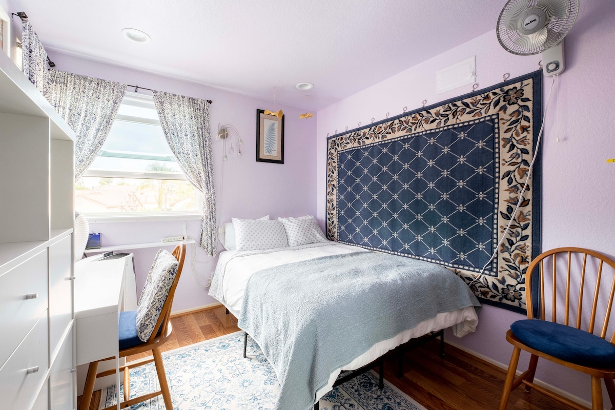 A serene bedroom is presented, featuring light purple walls and natural wood flooring. A spacious bed is adorned with soft, layered bedding. A desk and chair are positioned beside a window, with decorative curtains framing the view. An elegant tapestry is displayed on the wall, adding visual interest.