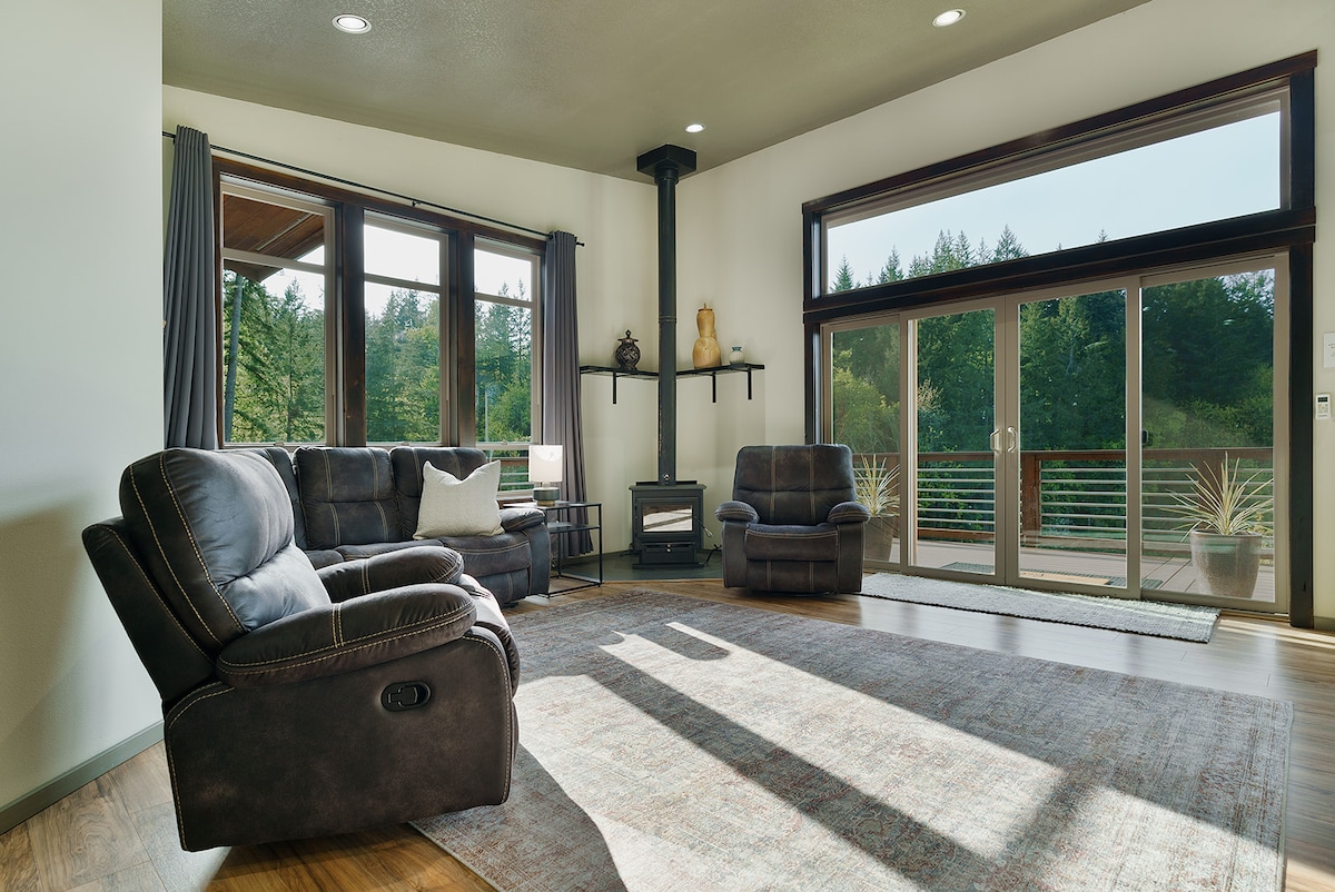 A spacious living area features comfortable reclining couches positioned for relaxation. Large windows allow natural light to fill the room, offering views of the surrounding greenery. A wood stove adds warmth to the space, complemented by a subtle area rug and minimalist decor.
