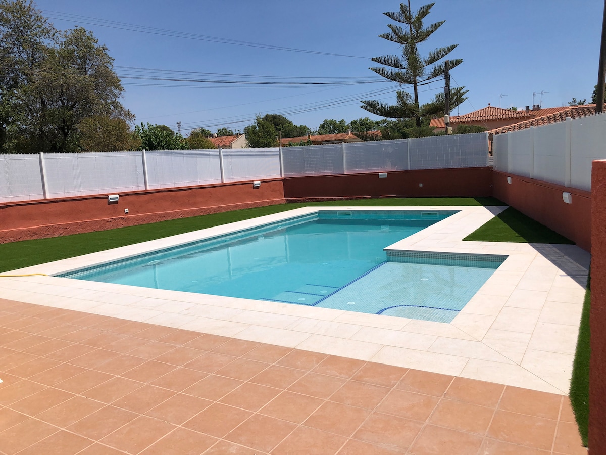 A spacious outdoor pool is surrounded by a newly paved deck with beige tiles. Lush green grass borders the area, creating a welcoming contrast. Palm trees are visible in the background, alongside a white fence that provides privacy.