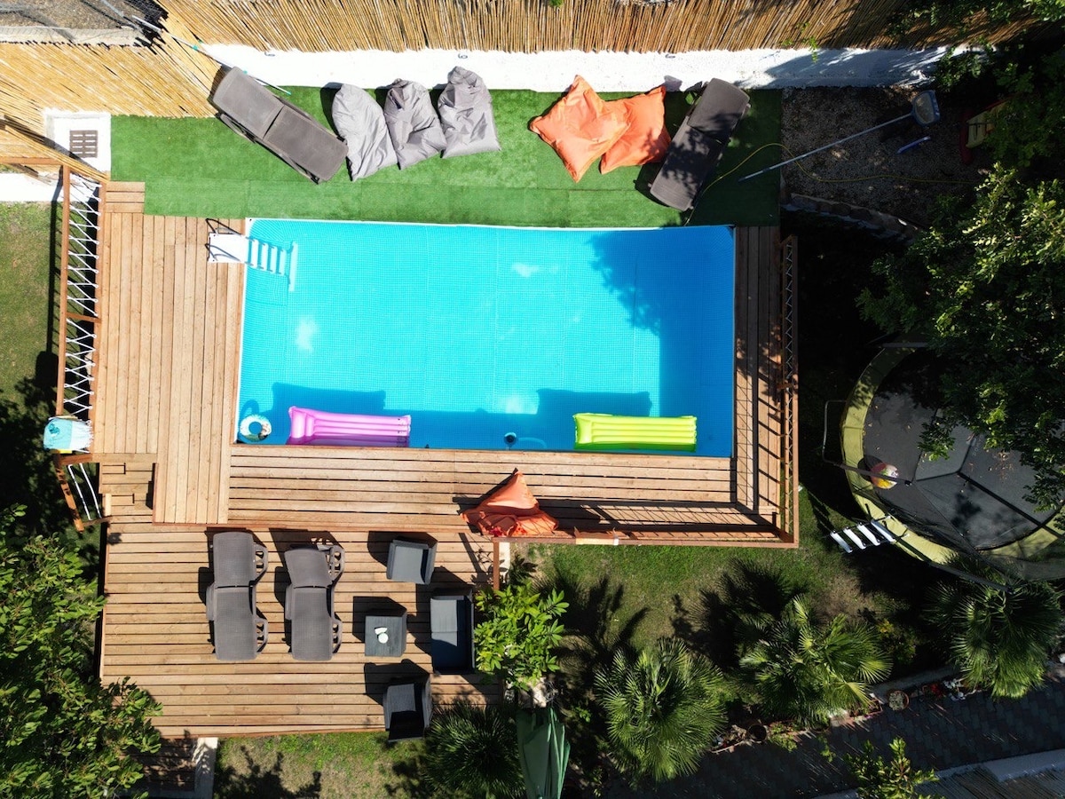 An aerial view reveals a rectangular swimming pool surrounded by wooden decking. Lounge chairs and bean bags in various colors are arranged on the grass, inviting relaxation. Tropical greenery complements the outdoor space, providing a serene atmosphere for guests to enjoy.