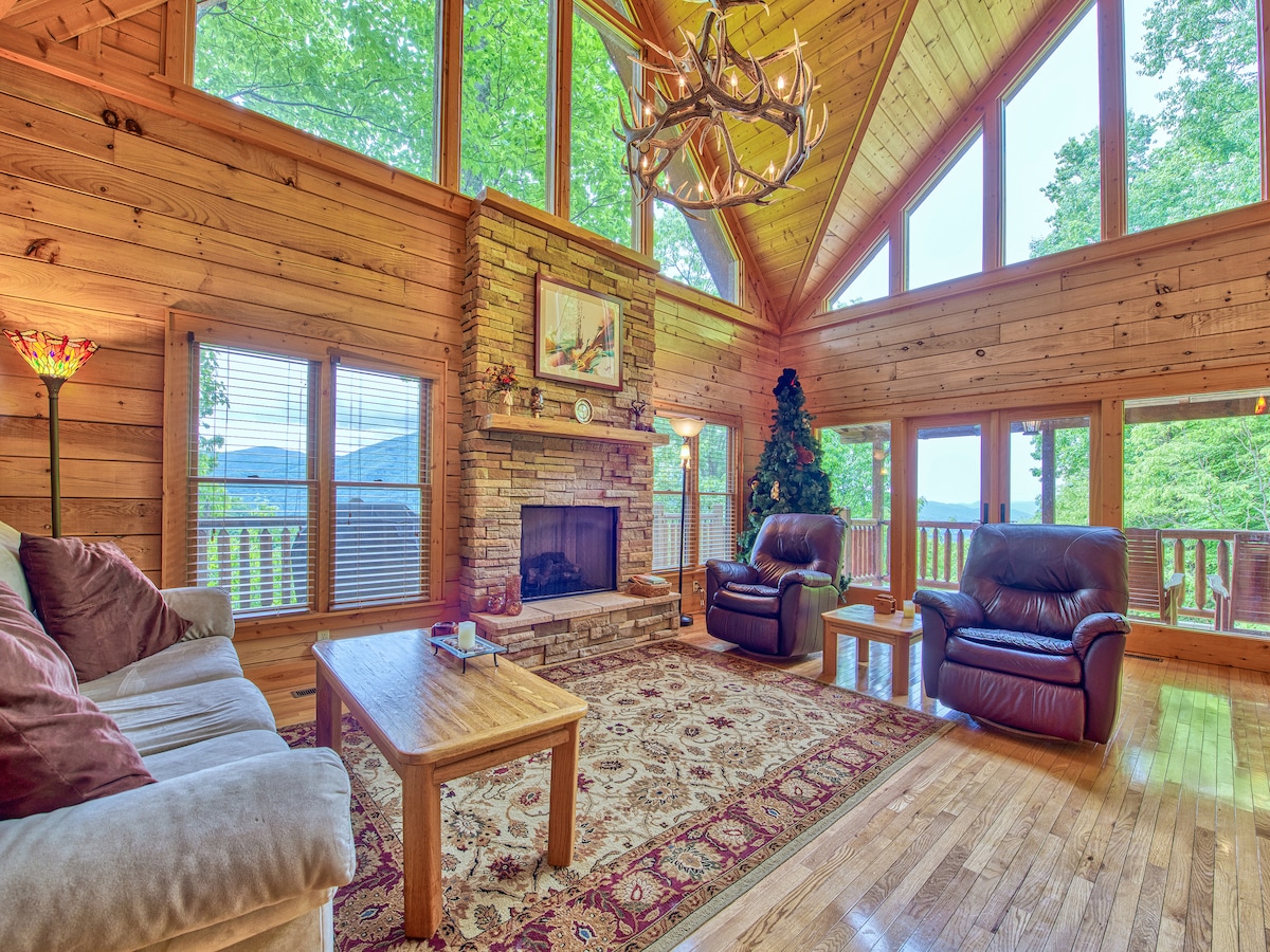 A spacious living room features floor-to-ceiling windows that provide expansive mountain views. Wooden walls and high ceilings create a warm ambiance. Two leather recliners and a sofa surround a central coffee table, while a stone fireplace adds a cozy focal point. A large area rug enhances the inviting atmosphere.
