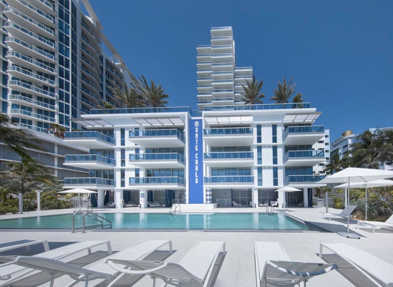 The luxury Monte Carlo building is depicted, featuring a modern architectural design with clean lines. A clear swimming pool is situated in the foreground, surrounded by white lounge chairs. Palm trees and tall buildings create a vibrant coastal atmosphere under a bright blue sky.