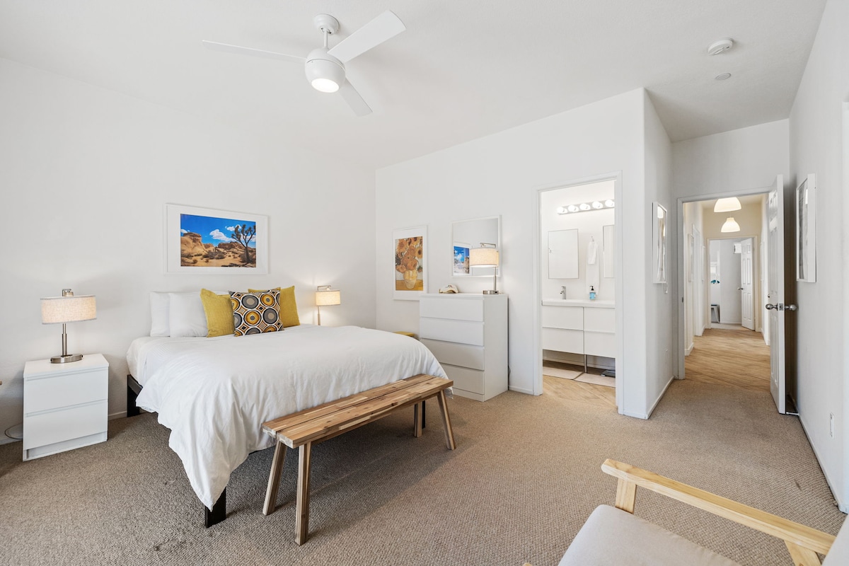 A spacious main suite is seen with neutral tones. A large bed with a white duvet is complemented by decorative pillows. Bedside tables with lamps are present, and a wooden bench provides additional seating. Soft lighting illuminates the room, highlighting artworks on the wall.