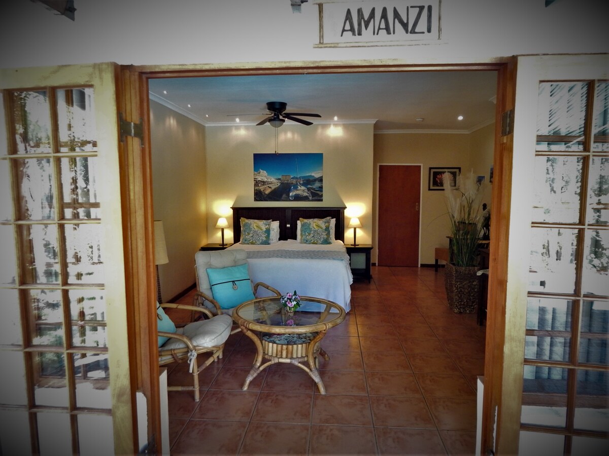 A view into a welcoming guest room is presented, featuring a spacious bed with decorative pillows, a cozy seating area with rattan furniture, and a soft color palette. French doors open to create an airy atmosphere, while soft lighting enhances the inviting space.