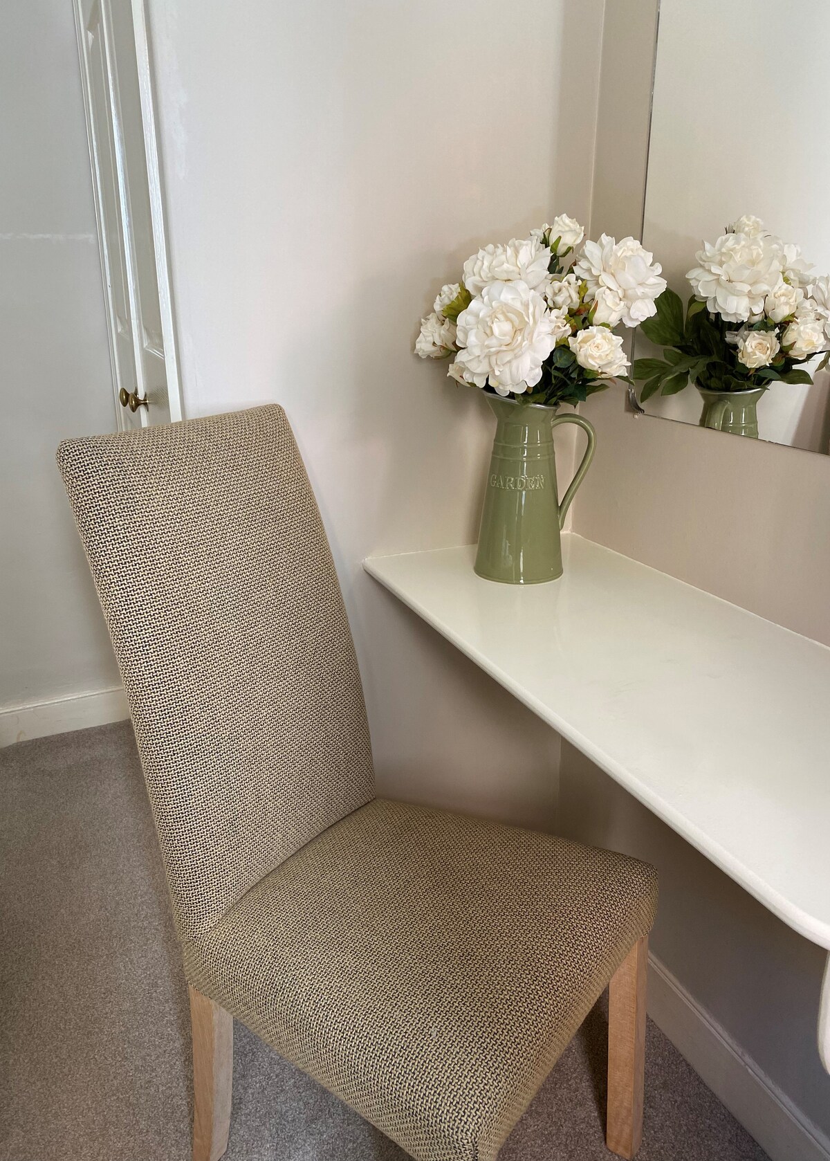 A simple dressing table is positioned against a light-colored wall. It is accompanied by a textured chair, and a vase filled with white flowers adds a touch of decor. A mirror above the table reflects the serene setting.