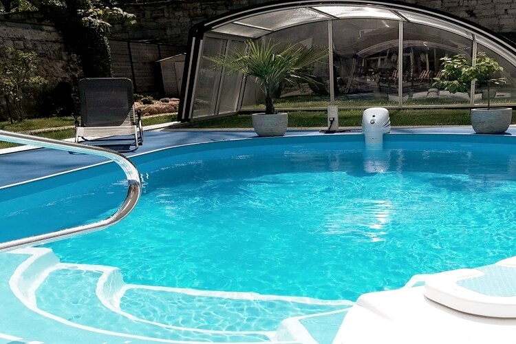 A heated, covered swimming pool is visible, surrounded by a well-maintained garden. Clear blue water sparkles under natural light, with a lounge chair positioned nearby. The pool is accessible and designed for relaxation in a tranquil outdoor setting.
