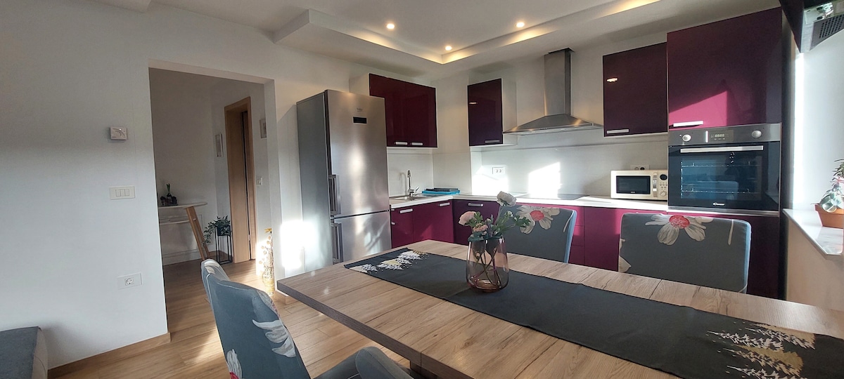 A modern kitchen space features burgundy cabinetry paired with stainless steel appliances, including a large refrigerator. A dining table with six chairs is set for meals, complemented by a decorative floral centerpiece. Natural light fills the area through a window, enhancing the inviting atmosphere.