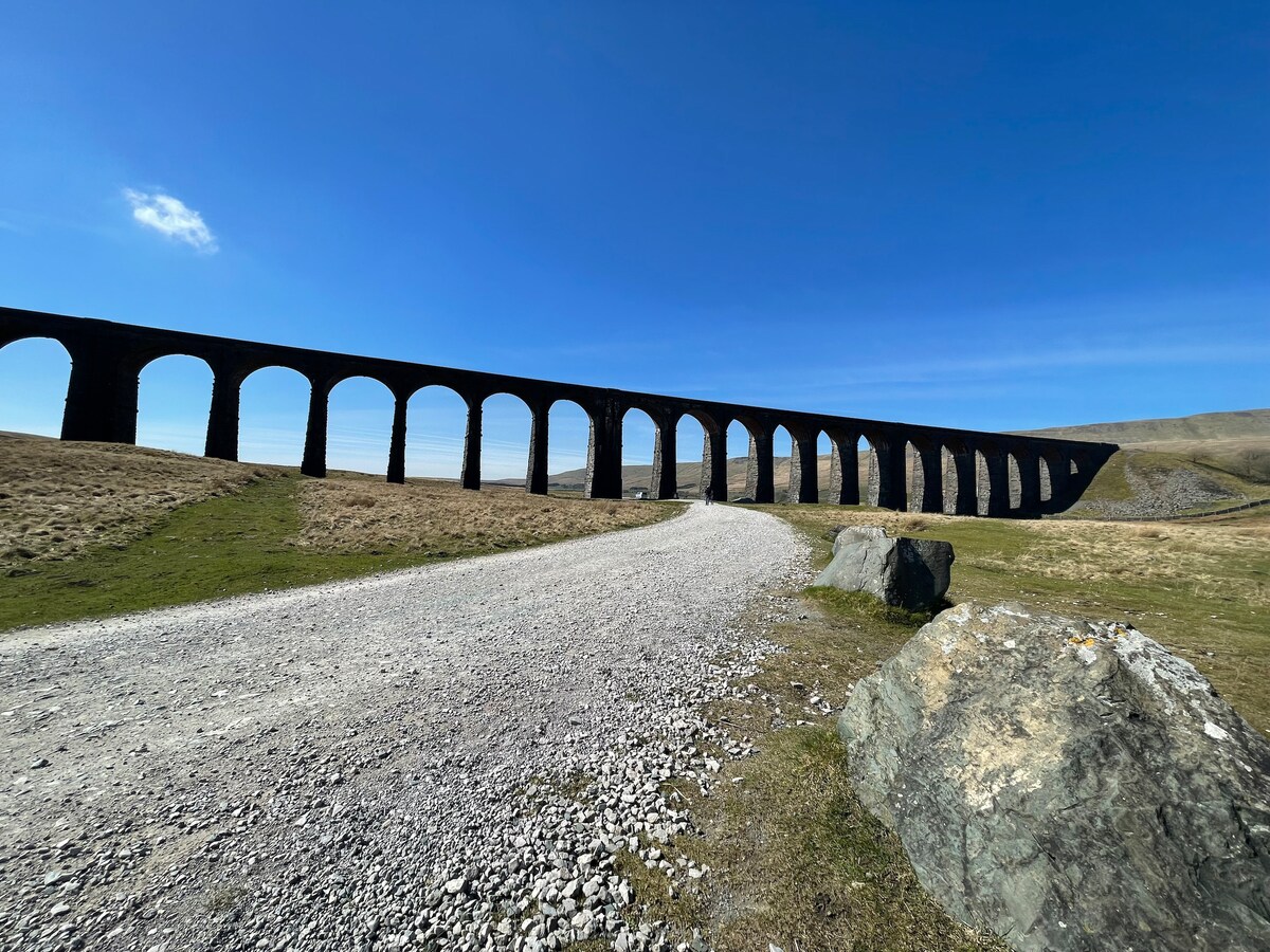 An expansive view of an impressive stone viaduct is presented, stretching across the landscape. The structure features multiple arches supported by sturdy columns, set against a backdrop of blue sky and rolling hills. A gravel path leads towards the viaduct from the foreground.