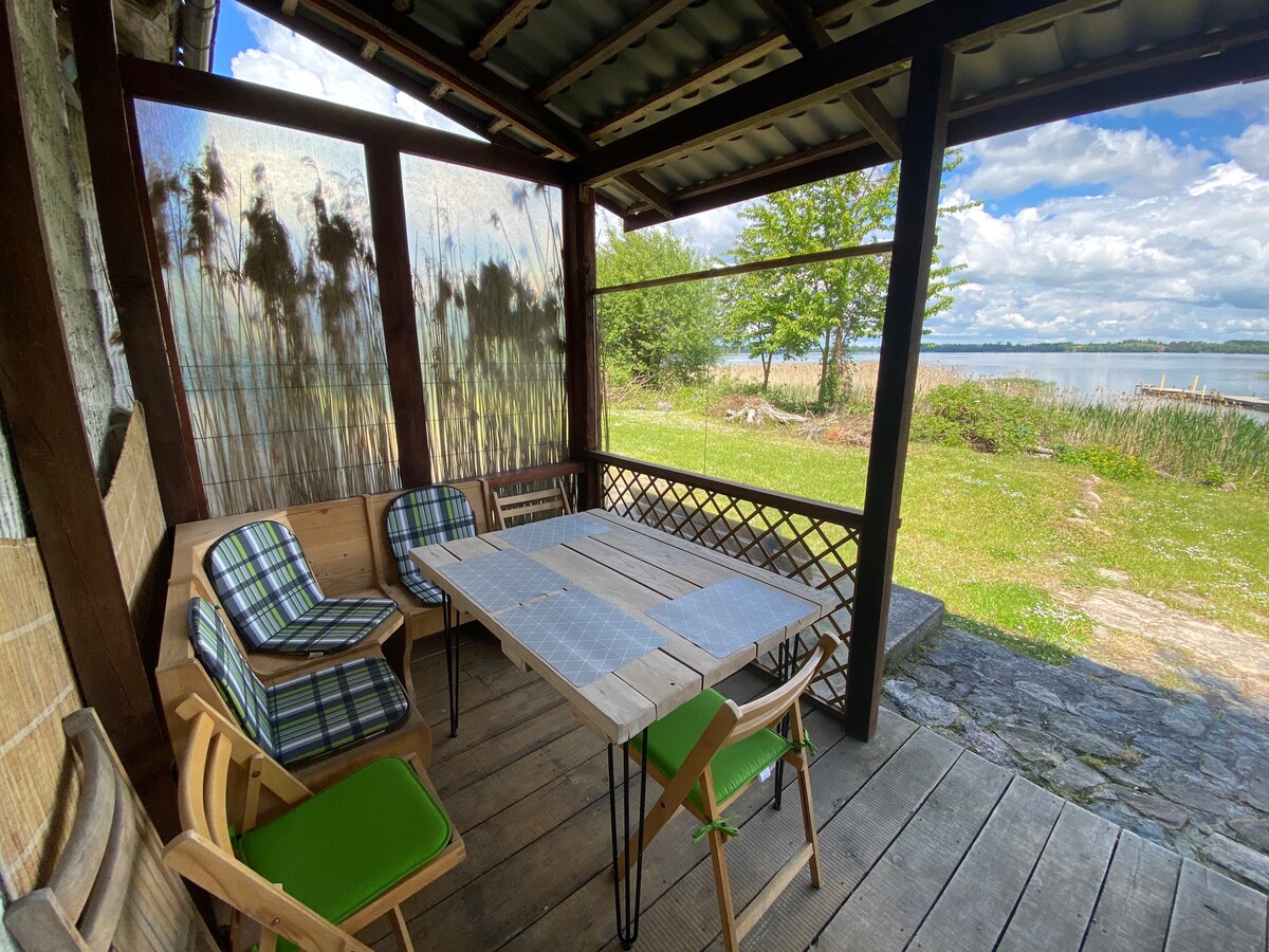 A wooden terrace overlooks the serene lake, furnished with a large table and four chairs. Natural light filters through the screen, highlighting grass and reeds in the vicinity. The calm water and greenery contribute to a peaceful setting for outdoor dining.