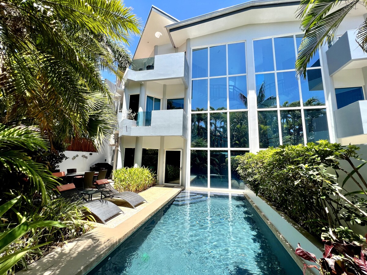 The villa is surrounded by lush tropical greenery, featuring a private pool with crystal-clear water. Expansive glass windows create a bright, airy atmosphere, while comfortable outdoor seating is arranged near the pool, offering a serene space for relaxation.