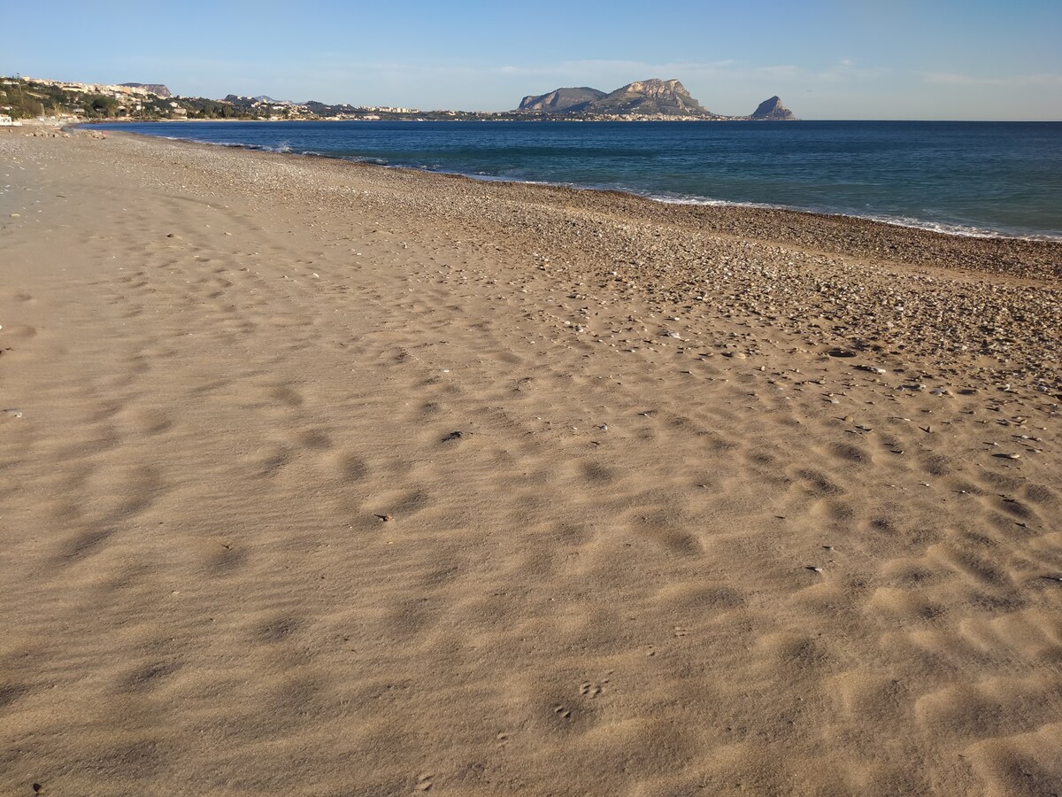 A sandy beach stretches along the shoreline, with smooth textures and patterns in the sand. The tranquil sea reflects clear blue skies, while distant hills are seen along the horizon, providing a serene backdrop to this coastal scene.