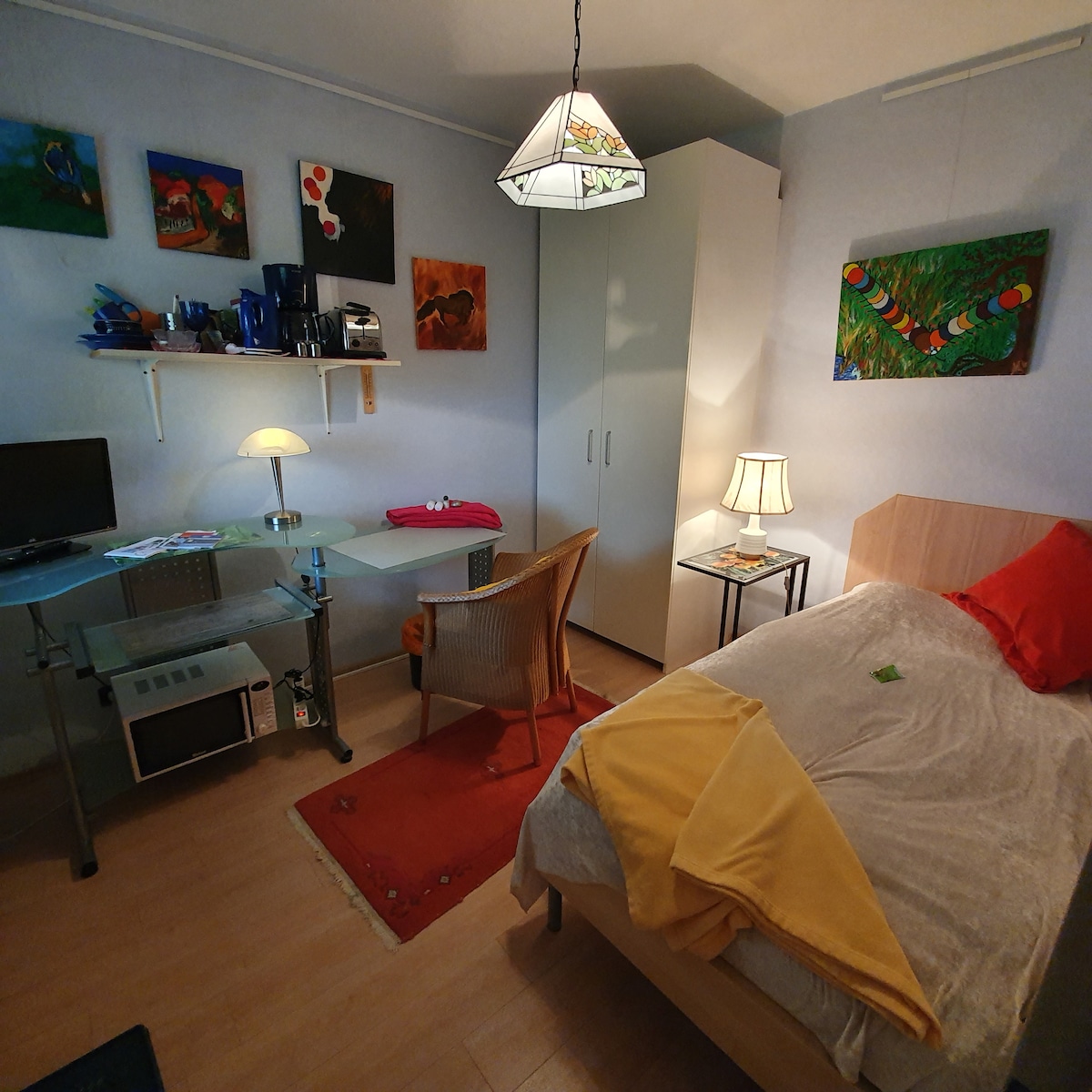A modern room features a spacious bed with a light-colored blanket and vibrant red accent pillows. A glass desk is paired with a wooden chair. Colorful artwork adorns the walls. A lamp and a TV are visible, along with a wardrobe and a microwave in the corner.
