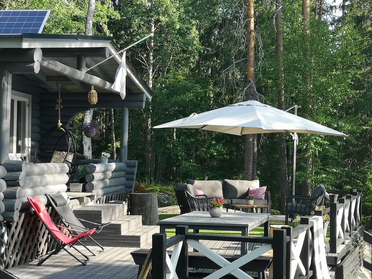 A wooden terrace is presented, featuring a large table surrounded by several seating areas. A shaded umbrella rests over the table. Comfortable lounge chairs and decorative plants enhance the relaxing atmosphere, while trees provide a green backdrop.