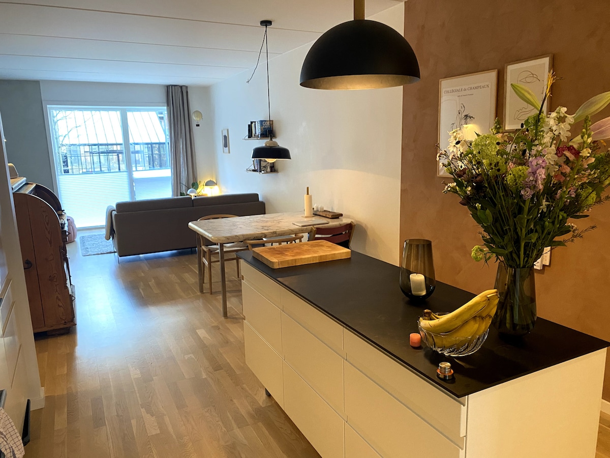 A modern kitchen area features a dark countertop with a bowl of bananas and a candle. A dining table is visible, and a comfortable sofa is placed across from it. Light enters through a large window, highlighting the warm tones of the wall.