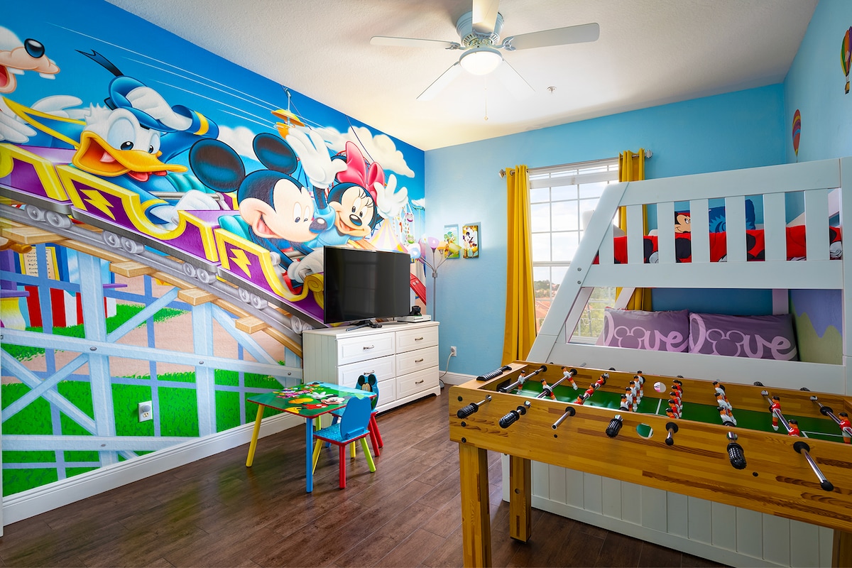 A vibrant children's bedroom is designed with a colorful mural featuring Disney characters. The room includes a bunk bed, a foosball table, and a television. Natural light is provided through a window, enhanced by cheerful yellow curtains. A small table with chairs is also present.